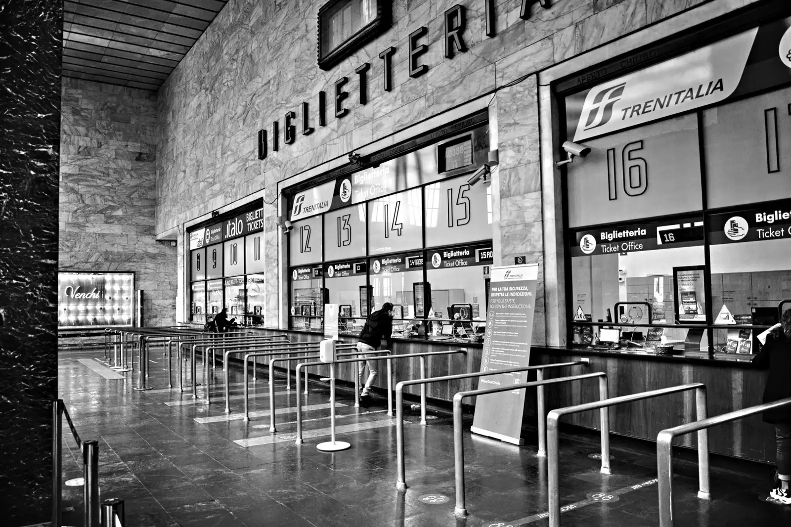 Monochrome view of an indoor train ticket area with counters and queue barriers.