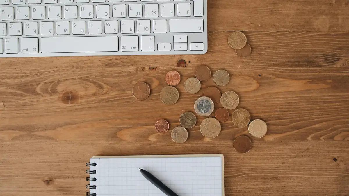 Overhead view of scattered coins on a weathered wooden desk next to a keyboard and open notebook for realistic travel budget.